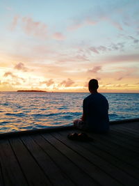 Rear view of man sitting on shore against sunset sky