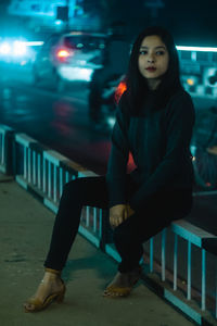 Portrait of young woman sitting on illuminated walkway at night