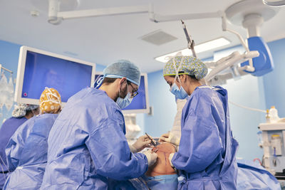 Doctors wearing face mask operating surgery while standing with colleague in background at operation room