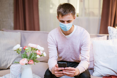 A young man sits on a sofa wearing a protective mask with a phone in his hand