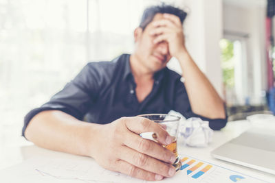 Depressed businessman holding whiskey glass while sitting with head in hands at office desk