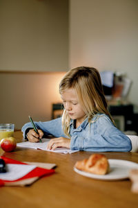 Focused boy with blond long hair writing on book while dong homework at home