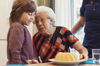 Senior woman talking to great grandson looking at sponge cake on table with caretaker standing at home