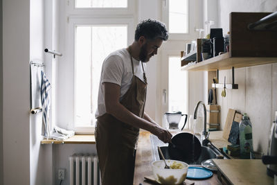 Side view of man wearing apron washing dishes in kitchen at home
