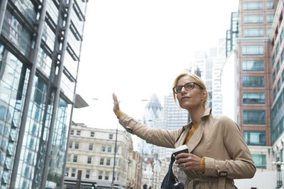 Woman with coffee cup waving hand while standing in city