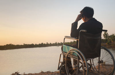 Rear view of man sitting by lake against sky during sunset