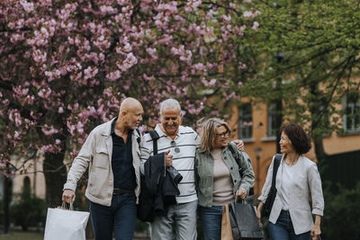 Happy male and female senior friends with shopping bags while in front of tree