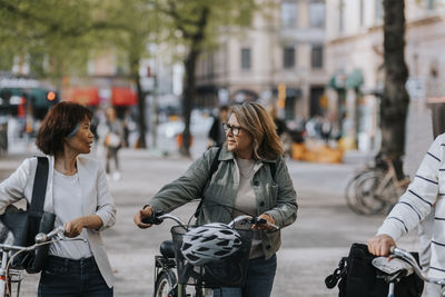 Senior female friends talking to each other while wheeling bicycles