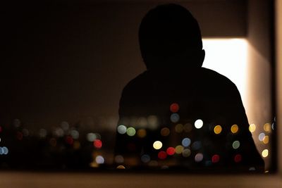Silhouette person by glass window with reflection of defocused lights