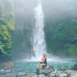 Full length of mid adult woman wearing bikini sitting on rock against waterfall