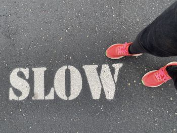 Low section of person standing on road in red sneakers by painted words that say slow