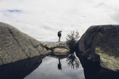 Rear view of hiker standing on rock by lake against cloudy sky