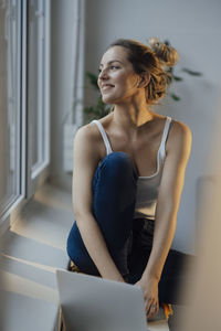 Smiling freelancer with laptop sitting on window sill at home