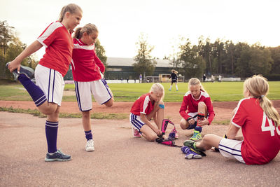 Girls stretching legs by friends wearing shin guards on field