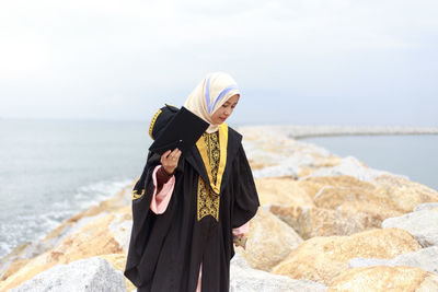 Young woman in graduation gown standing at beach against cloudy sky