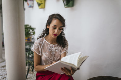 Content ethnic female in stylish outfit sitting on stool in patio and reading novel in book while enjoying summer weekend