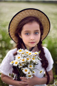 Portrait girl child in a white dress stands on a camomile field in a hat and with bouquet of flowers