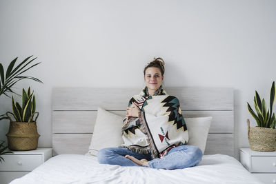 Young woman wrapped in shawl smiling while sitting on bed at home