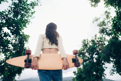 Rear view of woman holding skateboard while standing against trees