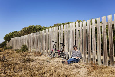 Woman reading book while resting against wooden fence