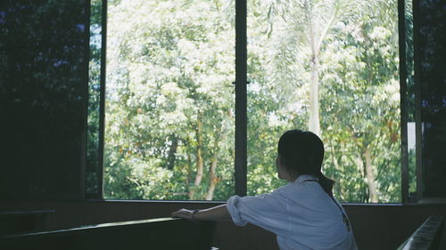 Woman looking trees through window while sitting on bench