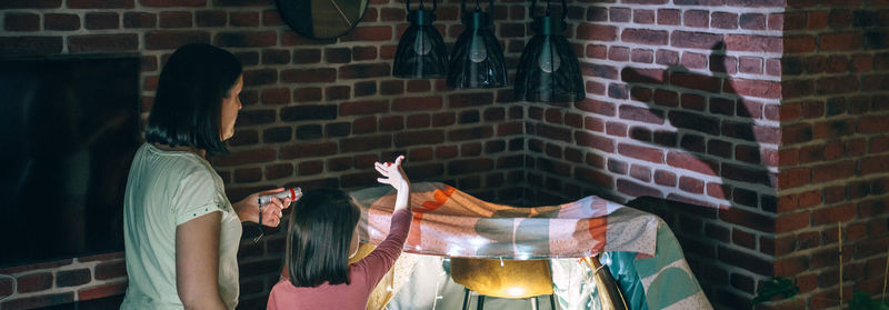 Banner of mother holding flashlight while daughter making shadow puppets with hands over wall.