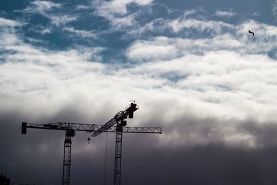 Silhouette cranes at construction site against cloudy sky