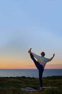 Rear view of woman with arms raised standing at beach against clear sky during sunset