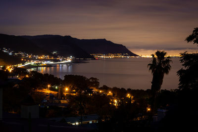 Illuminated city by sea against sky at sunset