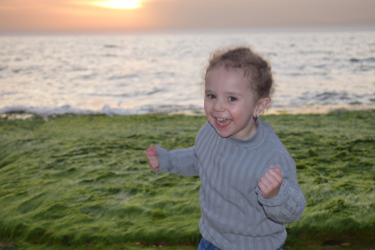 looking at camera, sea, boys, childhood, portrait, beach, waist up, one person, smiling, real people, happiness, casual clothing, elementary age, nature, focus on foreground, sunset, front view, leisure activity, standing, lifestyles, outdoors, water, horizon over water, sky, day, beauty in nature, people