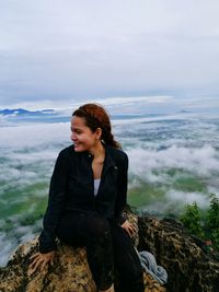 Smiling woman sitting on rock at mountain over clouds against sky