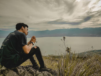 Side view of young man sitting against sky