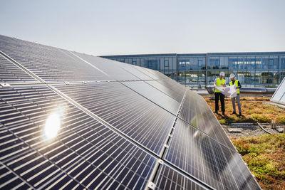 Two technicians analyzing blueprints on the rooftop of a corporate building equipped with solar panels