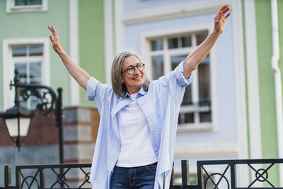 Portrait of young woman with arms raised standing against building