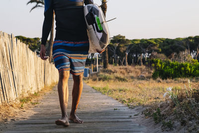 Unrecognizable surfer walks down the wooden walkway to the beach with a surfboard at sunrise.
