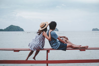 Rear view of couple sitting on railing by sea against sky