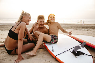Cheerful parents enjoying with daughter sitting on paddleboard at beach during sunset