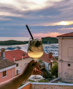 Woman holding cocktail, glass, personal perspective, sunset, sky in background.
