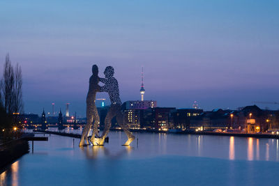 Molecule men sculpture on river in city against sky at night
