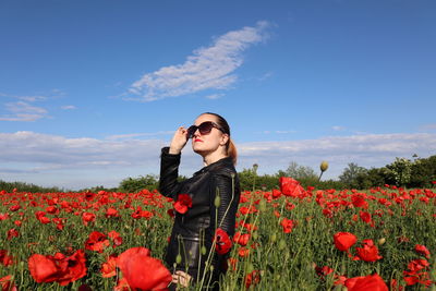 Young woman standing by poppy flowers on field against sky