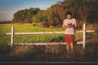 Mid adult man using mobile phone while standing by fence at farm