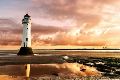 Lighthouse at beach against cloudy sky during sunset