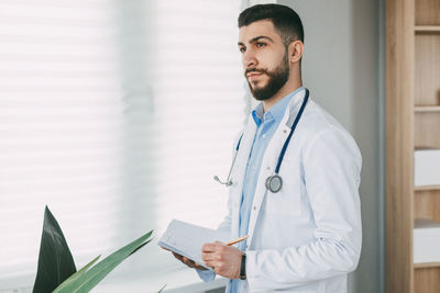 Modern young male doctor stands in his office and writes in his notebook