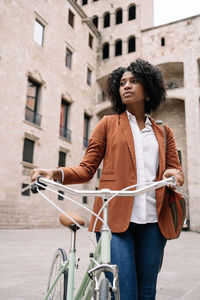 From below of black woman in smart casual style walking along street with bike and looking away