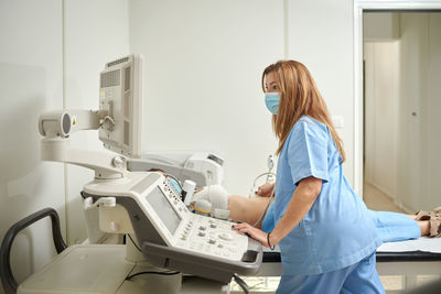 Side view of female doctor in uniform examining patient in mask while watching ultrasound scanner monitor in clinic