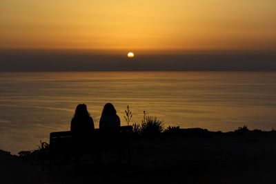 Silhouette women sitting on bench by sea during sunset