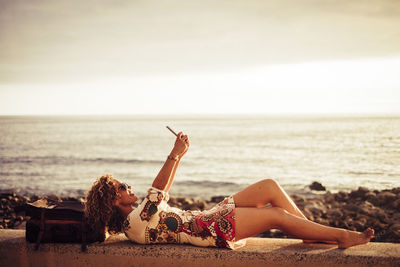 Side view of woman taking selfie while lying on retaining wall at beach against sky during sunset