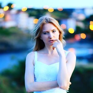 Portrait of young woman biting finger while standing in city during dusk