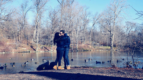 Kissing couple standing by lake against bare trees