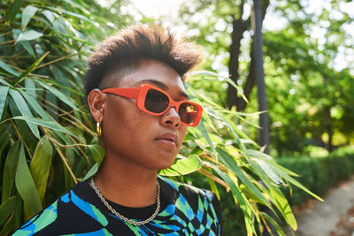 Filipino non binary person in stylish sunglasses with acne on face looking away while standing near tropical lush bush on summer day in park